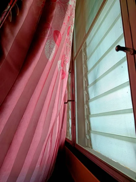 Soft pink curtain gently swaying beside a louvered window, capturing a serene morning breeze and warm natural light in a peaceful home interior
