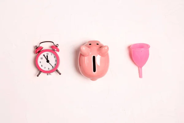 Menstrual cup with piggy bank and alarm clock on a white background. Save money with reusable feminine hygiene products.