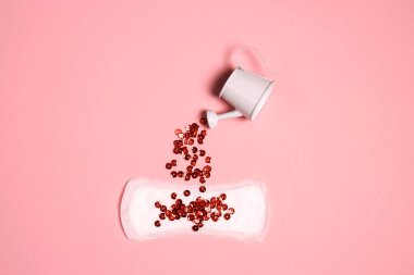 Kırmızı pullar küçük bir su kutusundan hijyenik peçeteye dökülüyor. Pembe arka plan. Adet dönemi, kadın hijyeni..
