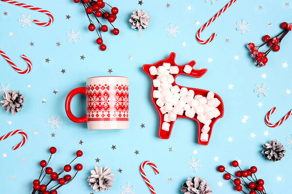 Winter mug with marshmallows in a plate in the form of a deer surrounded by christmas decorations on a blue background. Cozy festive hot drink with sweets.