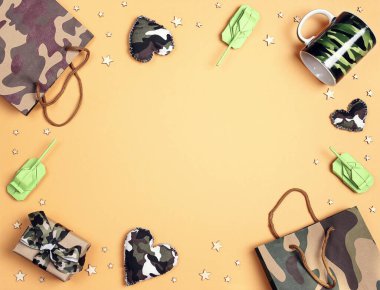 Kamuflaj kalpleri, hediyeler, çanta, kupa ve sarı arka planda origami kağıt tankları çerçevesi. Üst aşağı kompozisyon ve kop alanı. Anavatan Günü Savunucusu, 23 Şubat.