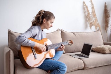 Evde, kanepede dizüstü bilgisayarın önünde gitar çalan genç bir kadın..