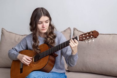 Uzun siyah saçlı bir genç evinde kanepede oturup gitar çalıyor..