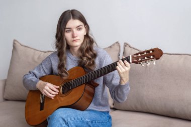 Uzun siyah saçlı bir genç evinde kanepede oturup gitar çalıyor..
