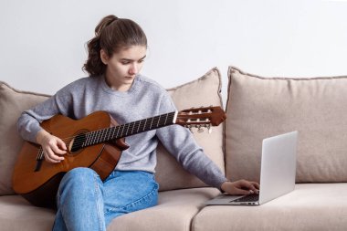 Evde, kanepede dizüstü bilgisayarın önünde gitar çalan genç bir kadın..