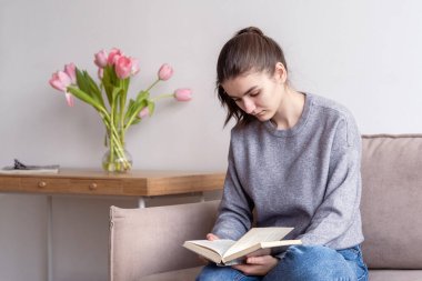 Bir genç evde kanepede oturmuş kitap okuyor..