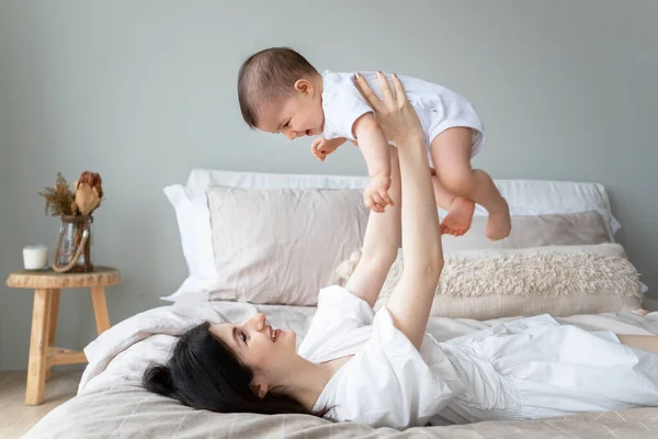 幸せな母親は赤ちゃんを空中に保持しています 幼い母親が子供と一緒にベッドで遊んでいる 幸せな子供時代 ストック写真