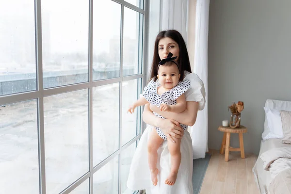 腕の中に9ヶ月の赤ちゃんを持つ若い母親が窓の近くに立っている 彼らはカメラを見て ストック画像