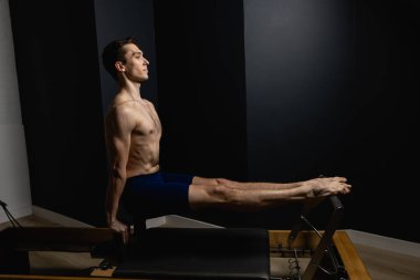 Sporty man doing exercises on pilates reformer training machine. Working out wearing sportswear, gym.