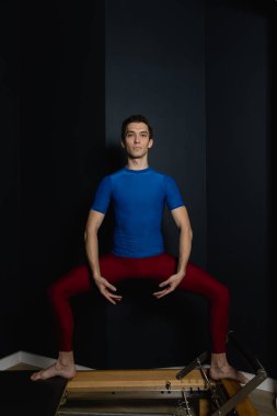 Sporty man doing exercises on pilates reformer training machine. Working out wearing sportswear, gym.