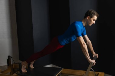 Sporty man doing exercises on pilates reformer training machine. Working out wearing sportswear, gym.