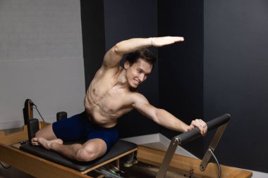 Sporty man doing exercises on pilates reformer training machine. Working out wearing sportswear, gym.