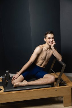 Sporty man doing exercises on pilates reformer training machine. Working out wearing sportswear, gym.