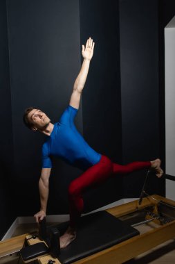Sporty man doing exercises on pilates reformer training machine. Working out wearing sportswear, gym.