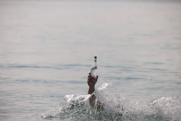 Bottiglia con acqua pura in mano in oceano e spruzzi intorno ad esso Foto Stock Royalty Free