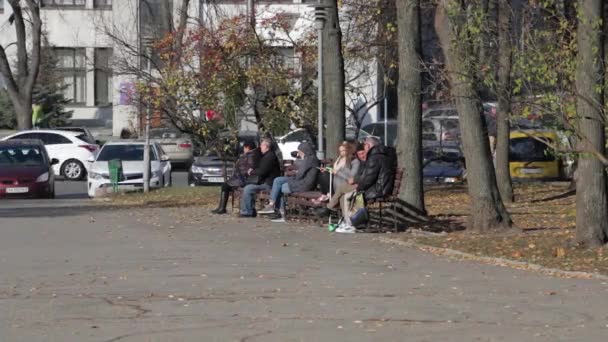 Unga och gamla människor sitter på en bänk på hösten nära en parkeringsplats. En ung flicka röker en cigarett i bakgrunden. — Stockvideo