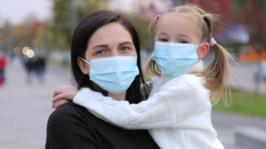 Şefkatli bir anne kızına mavi renkli cerrahi bir maske takarak sonbaharda soğuk hava koşullarından korunmak için kameraya bakar. Yavaş çekim yakın çekim