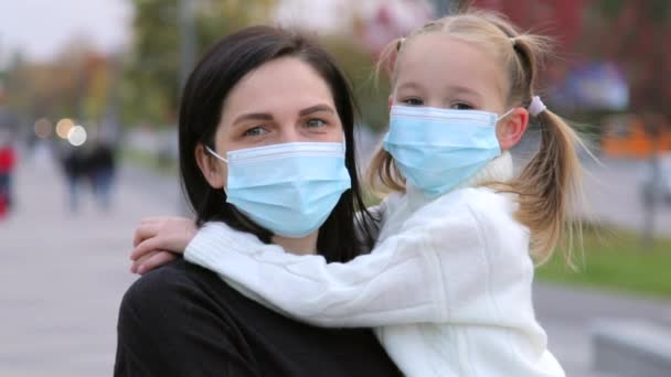 Una madre premurosa tiene la figlia tra le braccia in una maschera medica chirurgica di colore blu per proteggere contro le malattie infettive da un freddo all'aperto in autunno guardando la fotocamera. Primo piano al rallentatore — Video Stock