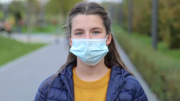 Ritratto di una bella ragazza caucasica affascinante in una maschera protettiva medica, guardando la fotocamera con attenzione. Donna di successo in posa per la fotocamera al rallentatore. Parco pubblico offuscato — Video Stock