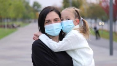 Siyah saçlı, beyaz bir kadının koruyucu bir tıbbi maske takmış kızıyla birlikte kameraya dikkatlice baktığı portresi. Kız annesine sıkıca sarılıyor ve ona yapışıyor. Hareket Görüntüsü