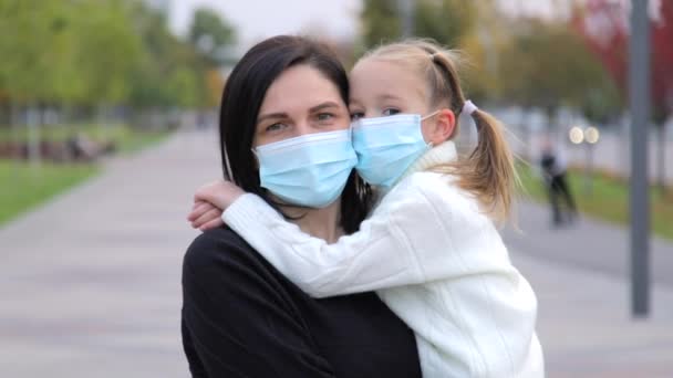 Retrato de una joven caucásica de cabello oscuro con su hija en una máscara médica protectora, mirando atentamente a la cámara. La niña abraza a su madre muy fuerte y se aferra a ella. Disparo de movimiento — Vídeos de Stock