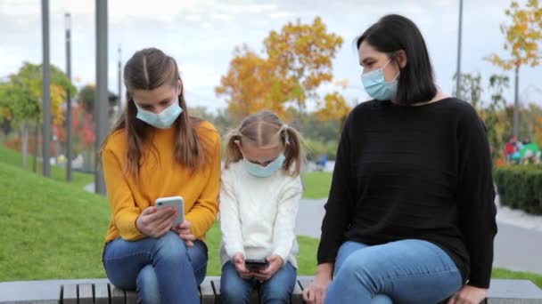 I bambini caucasici con una maschera medica protettiva fanno un gioco su un telefono cellulare mentre sono seduti su una panchina in un parco pubblico durante il giorno. Una madre premurosa porta via il telefono ai bambini perché — Video Stock