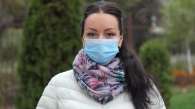 Sonbaharda sıcacık şık giysiler içinde sokakta duran ve çerçeveye bakan mavi koruyucu tıbbi maskeli, güzel, beyaz, esmer bir kızın yakın çekimi. Güvenli ve mutlu bir kadın