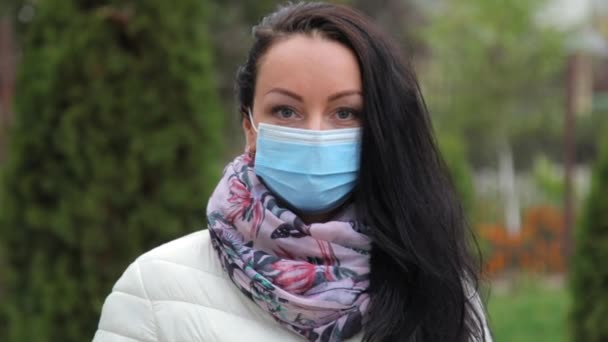 Close-up de uma bela jovem menina morena caucasiana em uma máscara médica de proteção azul que fica na rua com roupas quentes e elegantes no outono e olha para a moldura. Movimento lento. — Vídeo de Stock