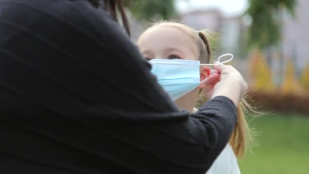 Una giovane madre caucasica si mette una maschera protettiva per sua figlia. La ragazza guarda sua madre e sorride. Sfondo parco sfocato primo piano rallentatore. — Video Stock