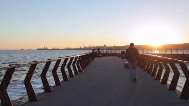 Sıcak sonbahar kıyafetleri içindeki yalnız bir erkek, Ukrayna 'nın Mariupol kentindeki Azov Denizi yakınındaki iskele boyunca gün batımında aktif olarak yürüyor. Yavaş çekim, nefes alan kamera..