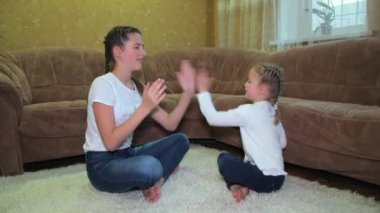 İki neşeli Kafkasyalı kız kardeş oturma odasındaki kanepenin yanında halıya otururken ellerini çırparak oynuyorlar. Ellerini kullanarak birlikte oyun oynayarak eğleniyorlar..