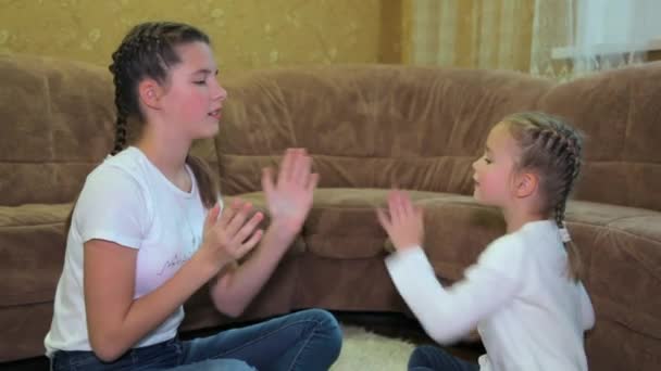 İki neşeli beyaz kız kardeş oturma odasındaki kanepenin yanındaki halıya otururken ellerini çırparak oynuyorlar. Ellerini kullanarak birlikte oyun oynayarak mutlu ve eğlenceli vakit geçiriyorlar.. — Stok video
