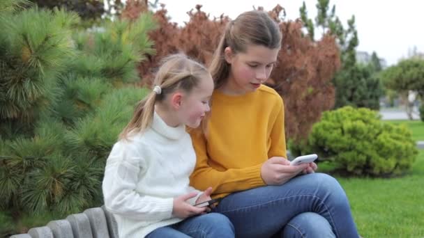 Hermanas niños usan teléfonos móviles fuera durante el día. Las niñas lindas se sientan en un banco en un parque público, miran un teléfono inteligente y se comunican. Modo vertical de teléfono móvil en dos manos. — Vídeos de Stock