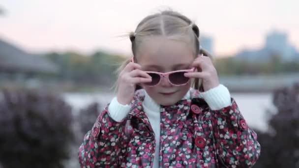 Портрет модной белой девочки в стильных солнцезащитных очках. Маленькая девочка стоит одна в городском общественном парке, смотрит в камеру и улыбается. Мода и стиль жизни. — стоковое видео