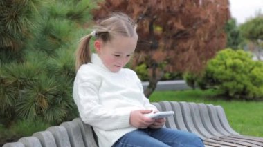 Güzel, küçük bir kız şehir parkında bir bankta tek başına oturuyor ve akıllı telefonuyla video oyunları oynuyor. Çocuk mutlu bir şekilde cep telefonunun dikey modunu iki elle kullanıyor..