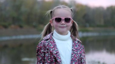 Modayı takip eden güneş gözlüklü hoş beyaz kız çocuğu. Şehir parkında tek başına olan küçük bir kızın portresi, kameraya bakar ve başparmağını kaldırır. Moda ve yaşam tarzı konsepti.