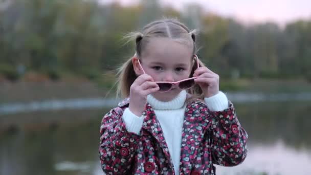 Portrait of a fashionable cute Caucasian child girl who is wearing stylish sunglasses. A little girl stands alone in a city public park, looks at the camera and smiles. Fashion and lifestyle concept. — Stok Video