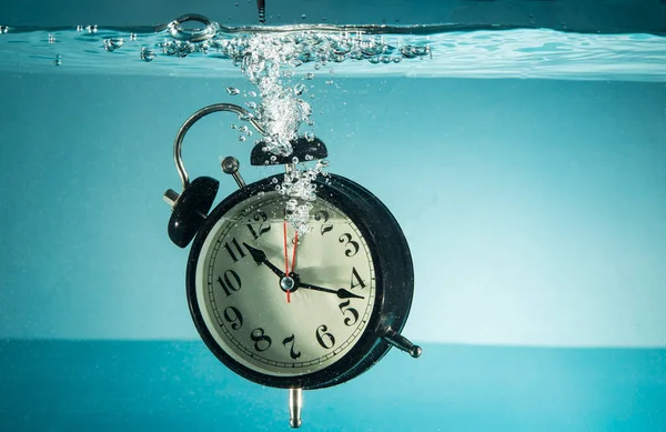 Table clock sinking into blue water tank - concept of time management with copy space.