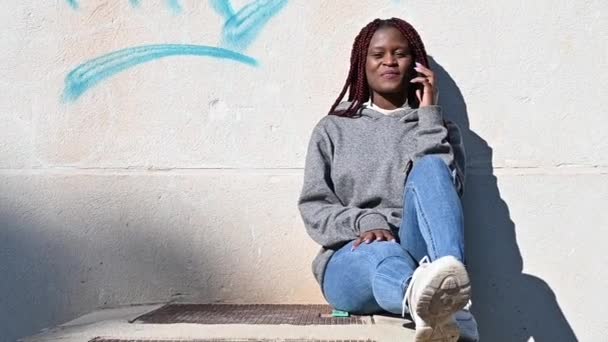 Νεαρή μαύρη γυναίκα κάθεται κοντά στον τσιμεντένιο τοίχο και μιλάει στο τηλέφωνο — Αρχείο Βίντεο