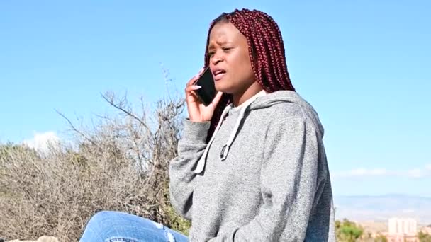 Молода чорна жінка сидить на одному камені і сперечається за допомогою телефону — стокове відео