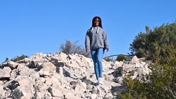 Africká Američanka sleze mezi kameny na modrém pozadí oblohy a usměje se — Stock video