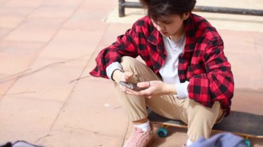 Akıllı telefondan yazı yazan genç bir kız kaykayın üzerinde oturuyor.