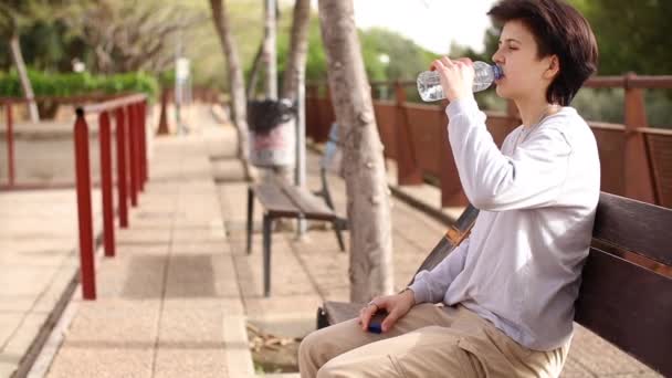 Chica adolescente cansada sentada en la silla y bebiendo agua de la botella — Vídeos de Stock