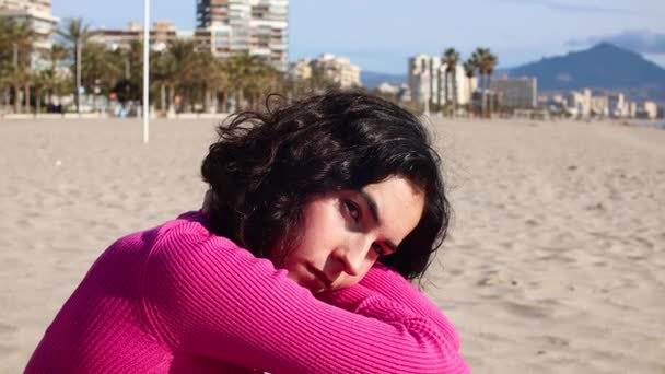 Sad girl in a pink sweater sits on the beach — ストック動画
