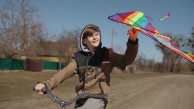 A happy boy with a flying rainbow kite rides a bicycle on the road.