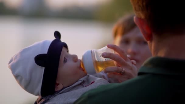 Молодые мама и папа кормят ребенка из бутылки в пруду на руках. — стоковое видео