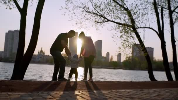 Dad and mom holding the hands of a child, look at the lake at sunset. — Vídeos de Stock