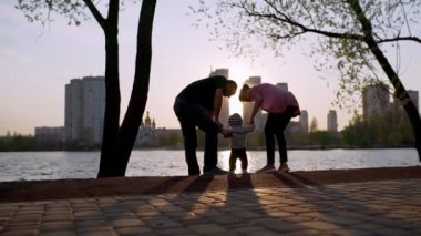 Anne ve baba bir çocuğun elini tutuyor, gün batımında göle bakıyorlar..