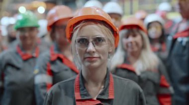 Bir kadın portresi, fabrika işçilerinin arka planına karşı kasketli ve gözlüklü bir endüstri işçisi. İşçi kameraya bakar. Endüstriyel işgücü kavramı.