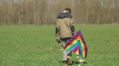 Bir çocuk elinde çok renkli bir uçurtma taşıyarak arazide yürür. Arkadan bak.